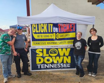 Students and an officer stand in front of Click It or Ticket and Slow Down Tennessee banners