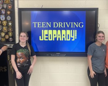 Students pose beside a Teen Driving Jeopardy game on a classroom screen